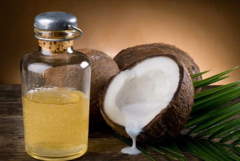 A bottle of coconut oil with a cork, split coconut, and palm leaves on a wooden surface