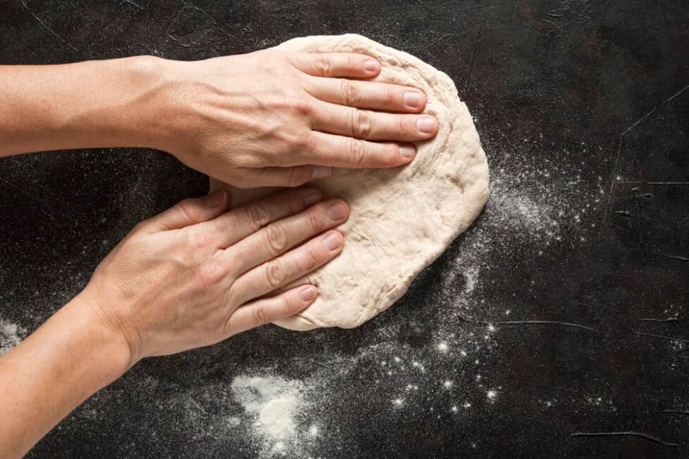 Hands flattening pizza dough on a floured surface