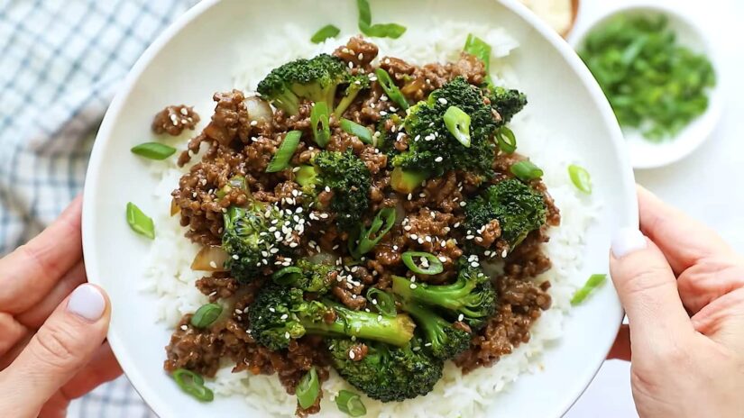 Hands holding a bowl of beef broccoli over rice, garnished with green onions